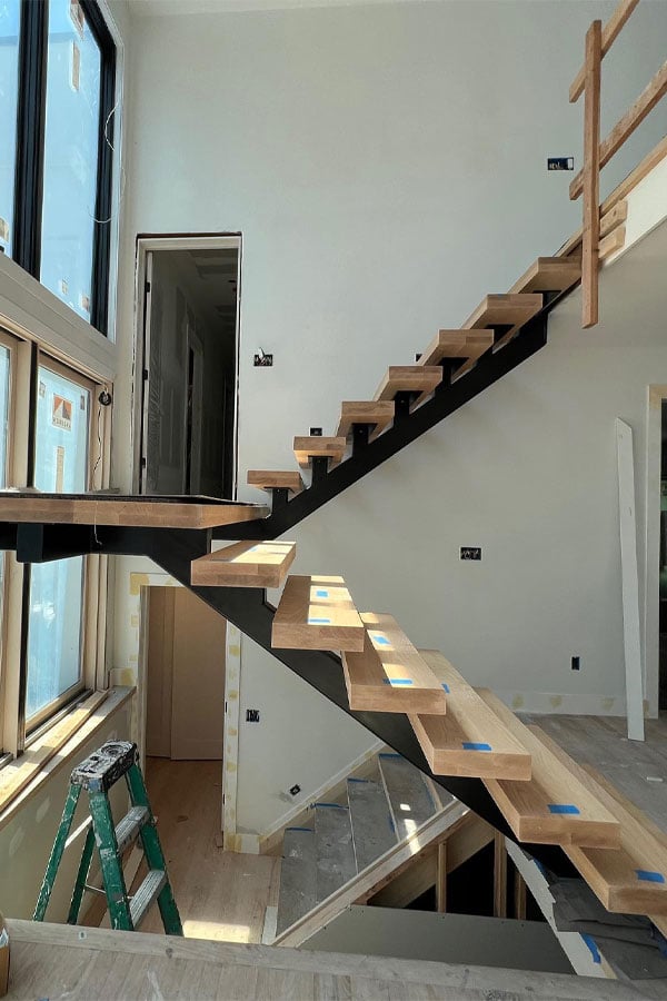 A staircase in process of being built faces a set of floor-to-ceiling windows.
