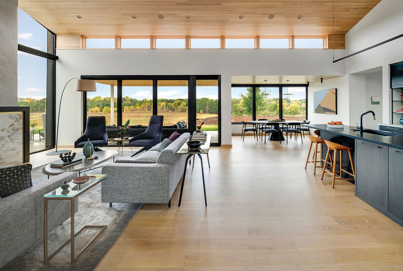 An open-concept space combining living room, kitchen, and dining room with views of a wetland and woods through the large black and natural wood windows.