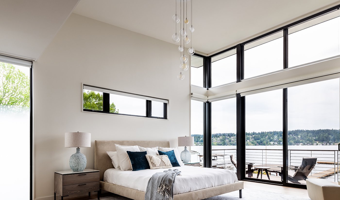 A bedroom with a wall of windows wrapping around a corner and a gliding patio door leading to a balcony with waterfront views. 