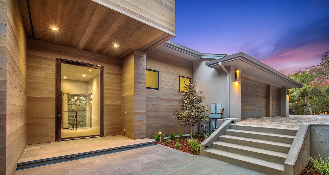 A large glass pivot door at the entrance of a modern home with wood siding and a two-door garage