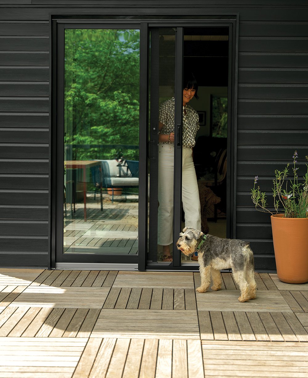 A patio with a dog and woman stepping through a gliding patio door with black frames. 