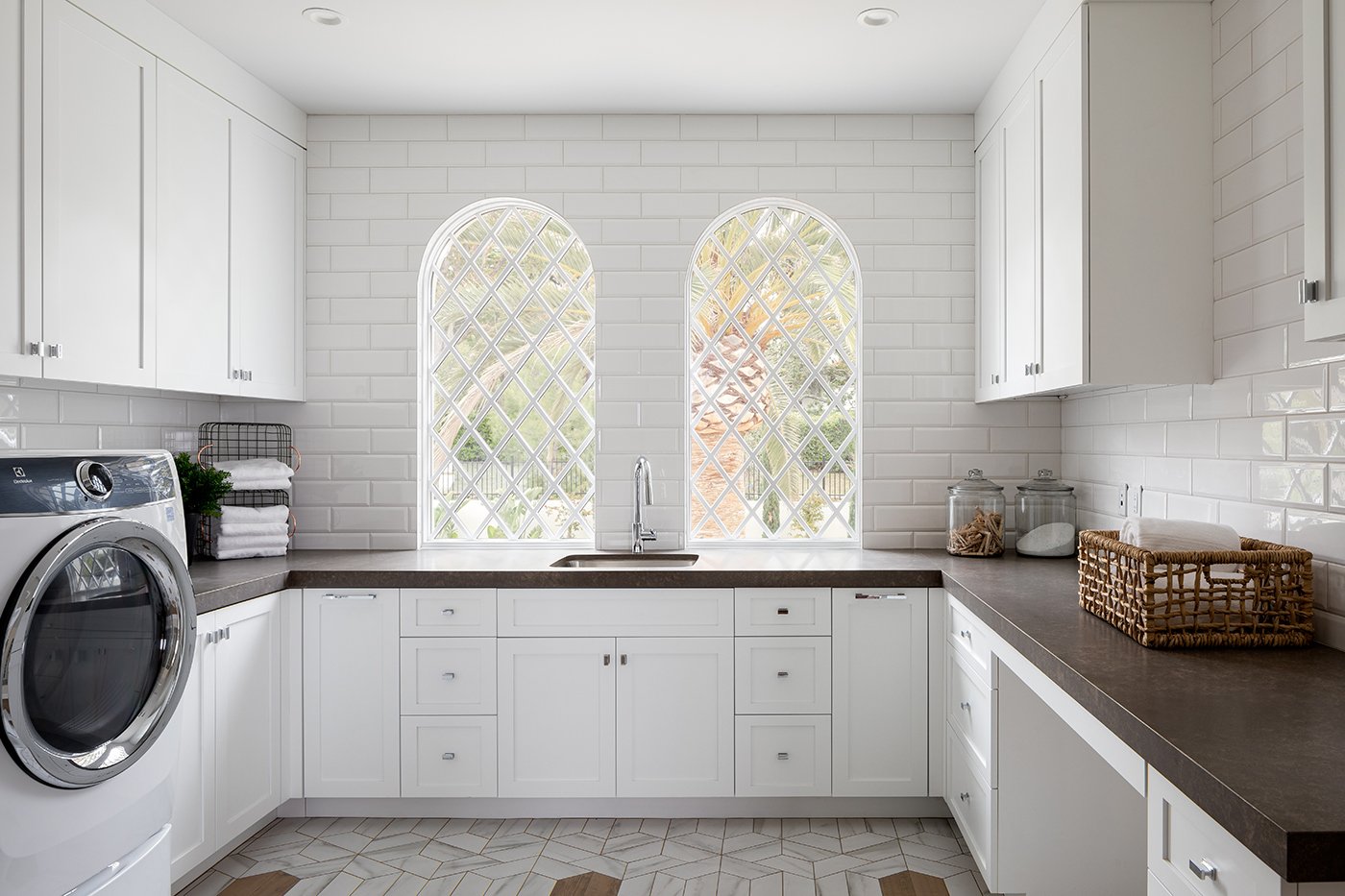 A laundry room with white cabinetry and tile and two Andersen® E-Series  Springline™ Windows behind the sink.