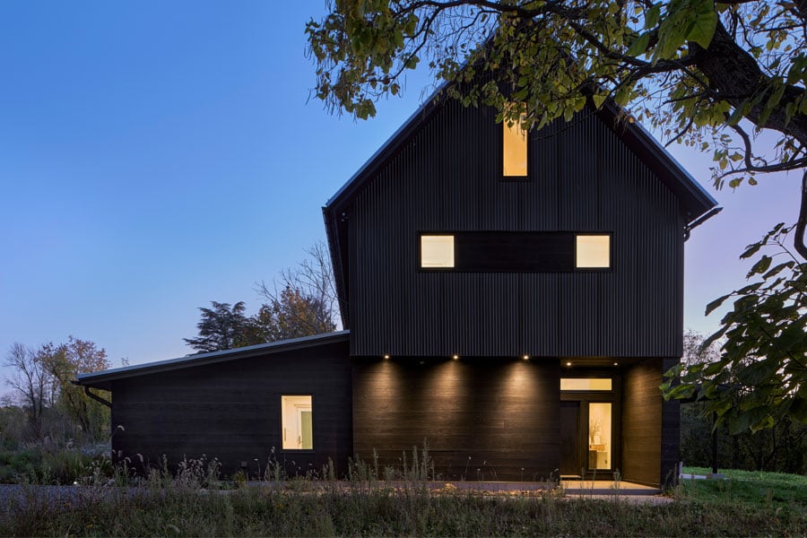 A twilight photo of a black barn-shaped home lit up on the interior.